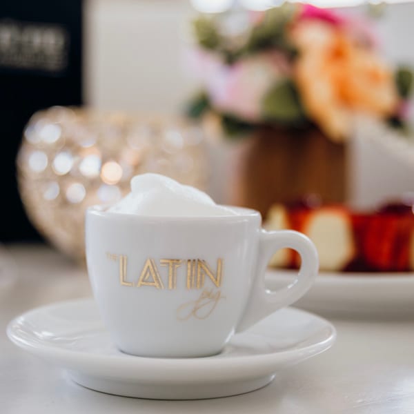 A white espresso cup with "The Latin Pig" logo, filled with frothy coffee, on a white saucer.