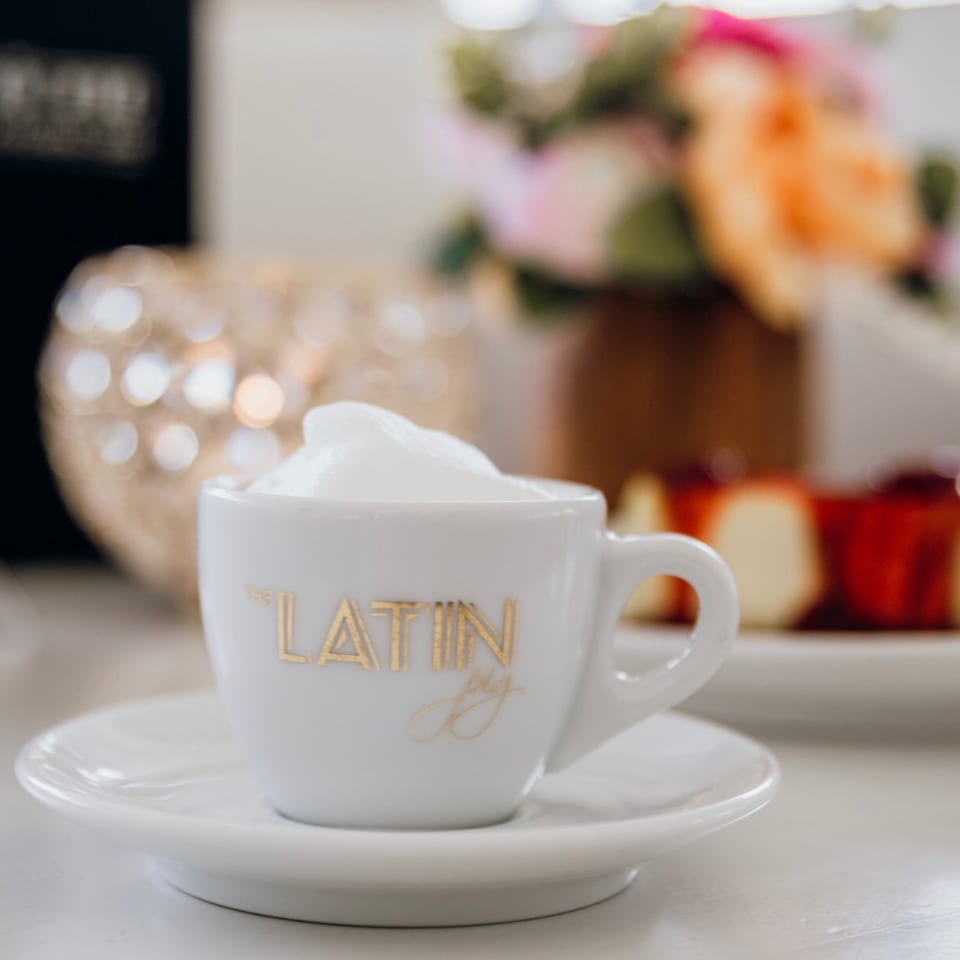 A white espresso cup with "The Latin Pig" logo, filled with frothy coffee, on a white saucer.