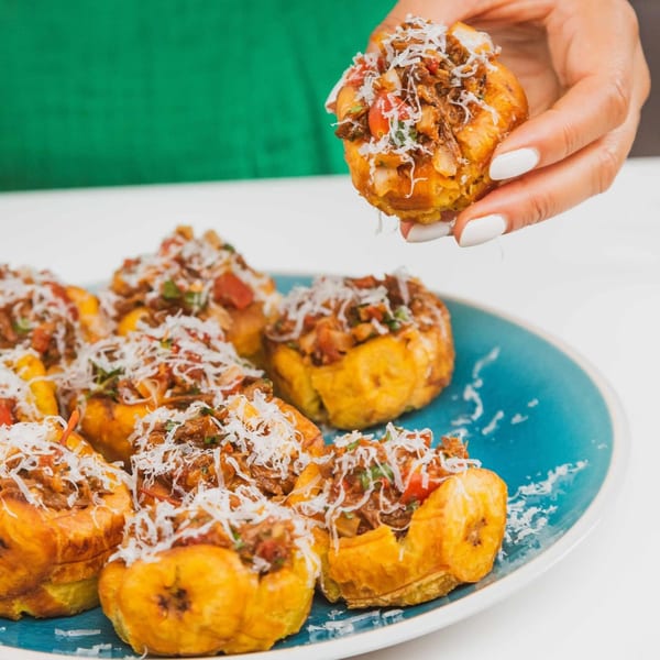 Hand holding a Cuban tostone cup filled with shredded meat and cheese, with more on a blue plate.