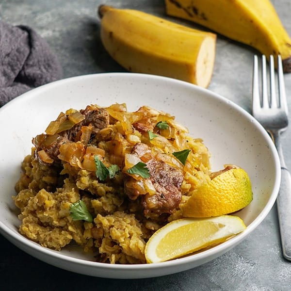 Cuban Mofongo with beef and onions, garnished with cilantro and lemon wedges, with plantains in the background.